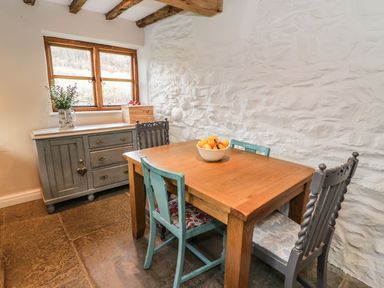 A dining area with a wooden table and four chairs next to a window and a cabinet with decorative items at Tan Y Garth Cottage in Glyndyfrdwy near Llangollen