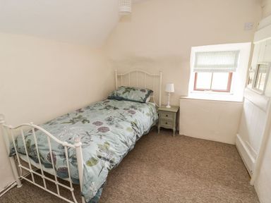A bedroom with a single bed floral bedding a nightstand with a lamp a window with a checked blind and a door at Tan Y Garth Cottage in Glyndyfrdwy near Llangollen