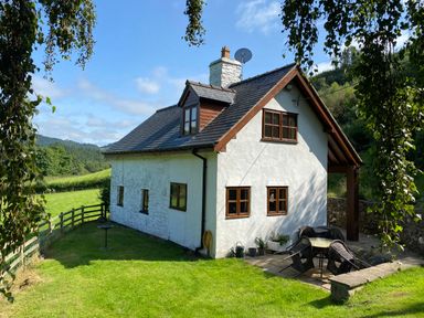 A white cottage with a slate roof and wooden windows in a green grassy area with outdoor seating at Tan Y Garth Cottage in Glyndyfrdwy near Llangollen