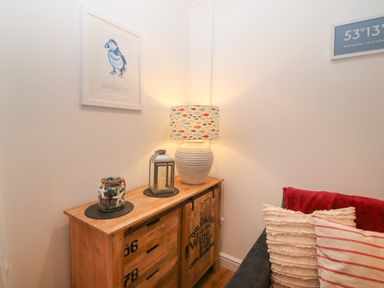 A corner with a wooden cabinet holding a fish patterned lamp a lantern and decorative jar with starfish above the cabinet is a framed puffin print next to a couch with cushions and a red throw at 8 Pen Llanw Tides Reach Rhosneigr