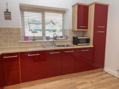 A kitchen with red cabinets a stainless steel sink a microwave and window with blinds at 8 Pen Llanw Tides Reach in Rhosneigr