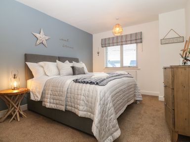 A bedroom with a bed with pillows and blankets a wooden side table with a lamp a window with blinds and a dresser at 8 Pen Llanw Tides Reach in Rhosneigr