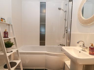 A bathroom with a bathtub shower combination a white sink a round mirror and a white shelving unit with toiletries and a plant at 8 Pen Llanw Tides Reach in Rhosneigr