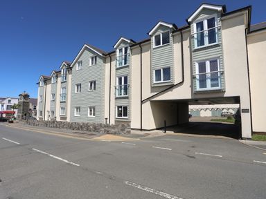 A street view of a multi-story residential building with light green and cream facades and an archway entrance at 8 Pen Llanw Tides Reach in Rhosneigr