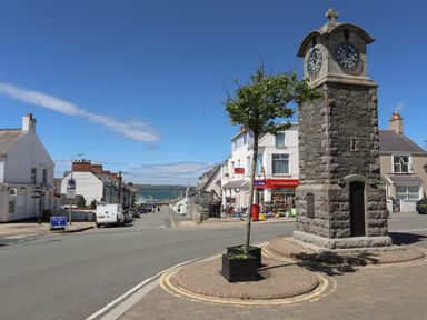 A street view with a stone clock tower, a tree in a planter, parked cars, shops, and buildings near the sea at 8 Pen Llanw Tides Reach in Rhosneigr