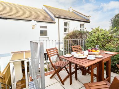 An outdoor patio with a wooden table and chairs set with plates of pastries cups and a bowl of fruit with a house and plants in the background at Little Tern Brewers Quay Harbour