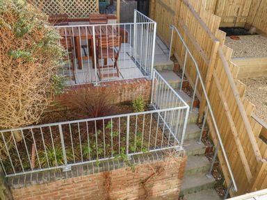 A backyard with a raised patio area with wooden dining furniture a small garden with plants and a staircase with metal railings at Little Tern Brewers Quay Harbour