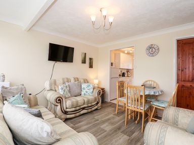 A living room with sofas a wall-mounted television a dining table with chairs and a kitchen in the background at Chalet 12 in Hemsby
