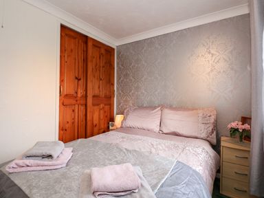 A bedroom with a bed covered in pink and gray bedding towels on the bed wooden closet doors and a nightstand with flowers at Chalet 12 in Hemsby