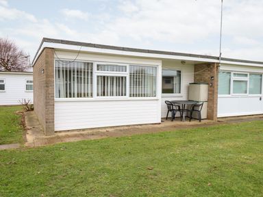 An exterior view of a single-story building with large windows a small patio with a table and two chairs and a grassy yard at Chalet 12 in Hemsby