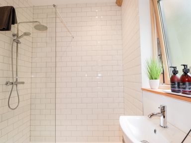 A bathroom with a shower area with white tiled walls a sink a window with toiletries and a small plant on the ledge at Pine Marten Lodge in Dulnain Bridge near Nethy Bridge