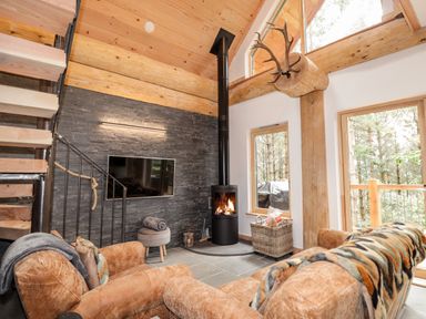 A living room with a wood burning stove a flat screen TV and a staircase with wooden beams and large windows at Pine Marten Lodge Dulnain Bridge near Nethy Bridge