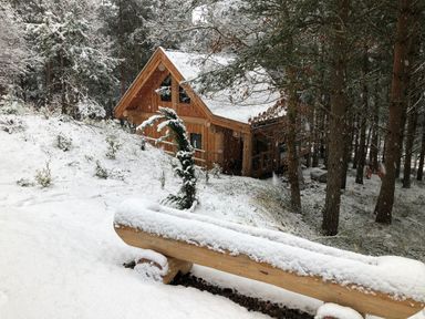 A wooden cabin in a snowy forest with a log bench covered in snow at Pine Marten Lodge Dulnain Bridge near Nethy Bridge