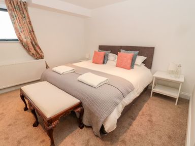 A bedroom with a double bed pillows and blankets a wooden bench bedside tables with lamps and a window with floral curtains at Shepherds House in Lowick