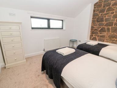 A bedroom with two single beds with dark blankets and towels on top a white chest of drawers a small round side table and a stone wall at Shepherds House in Lowick
