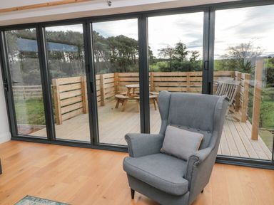 A gray armchair with a cushion near large sliding glass doors opening to a wooden deck with a picnic table at Shepherds House in Lowick