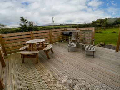 A wooden deck with a round picnic table with benches two wooden chairs and a barbecue grill at Shepherds House in Lowick