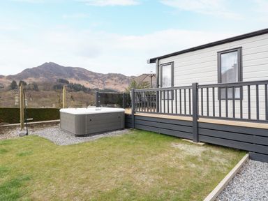 An outdoor area with a hot tub next to a deck attached to a white building with mountains in the background at Red Kite Lodge in St Fillans