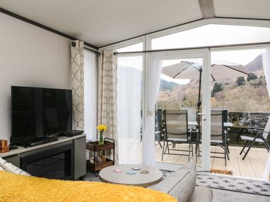 A living room with a television a round coffee table and sliding glass doors opening to a balcony with chairs and an umbrella at Red Kite Lodge in St Fillans