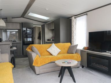A living room with yellow covers on sofas a round wooden coffee table and a TV with fireplace at Red Kite Lodge in St Fillans