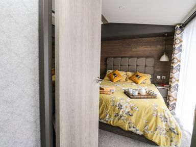 A bedroom with a bed with yellow floral bedding and pillows a wooden headboard bedside table with a lamp and curtains at Red Kite Lodge in St Fillans