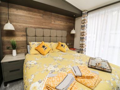 A bedroom with a bed with yellow floral bedding and bee pillows beside nightstands with lamps and a window with curtains at Red Kite Lodge in St Fillans