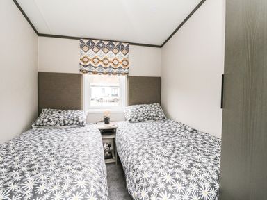 A bedroom with two single beds with daisy patterned bedding a window with a patterned blind and a small nightstand with a lamp and book at Red Kite Lodge in St Fillans
