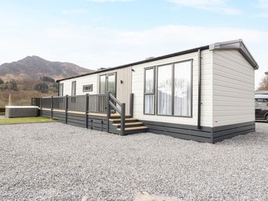 A mobile home with a deck and stairs on a gravel lot with mountains in the background at Red Kite Lodge in St Fillans
