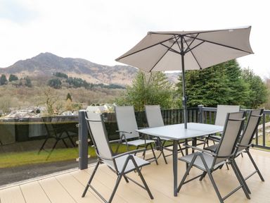 An outdoor patio with a table and six chairs under an umbrella with mountains in the background at Red Kite Lodge in St Fillans