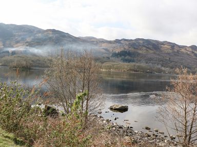 A lake with a rocky shore and leafless trees with hills in the background at Red Kite Lodge in St Fillans