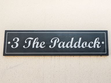 A black rectangular house sign with white text reading 3 The Paddock on a textured beige wall at 3 The Paddock in Benthall near Beadnell