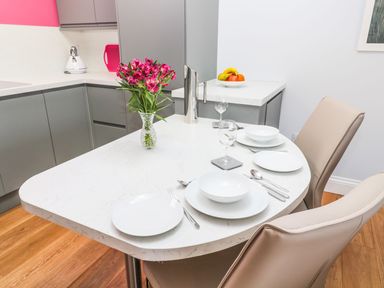 A kitchen dining area with a white countertop set with plates bowls silverware and wine glasses a vase of pink flowers and a fruit bowl at 3 The Paddock Benthall near Beadnell
