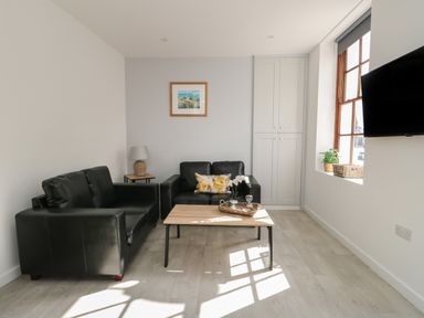 A living room with two black sofas a wooden coffee table a side table with a lamp a wall cabinet a window with plants and a wall-mounted TV at Llety Pen Deitsh 1 in Caernarfon