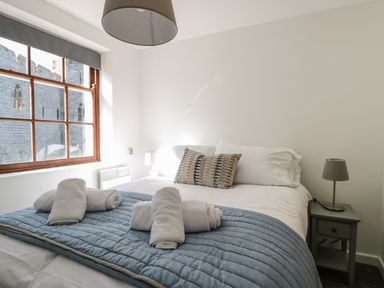 A bedroom with a bed with white linens and rolled towels near a window showing a stone building at Llety Pen Deitsh 1 in Caernarfon