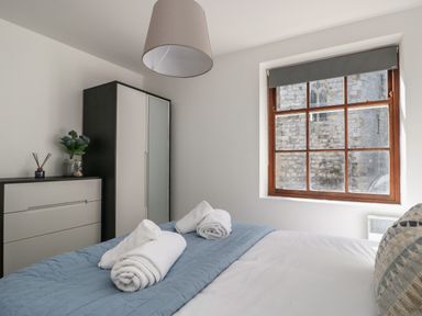A bedroom with a bed with rolled towels on a quilted blue cover and a window showing a stone building outside at Llety Pen Deitsh 1 in Caernarfon