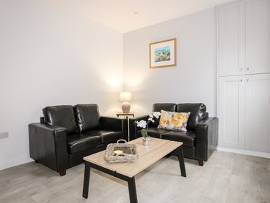 A living room with two black sofas a wooden coffee table with a wicker tray and flowers a lamp and wall art at Llety Pen Deitsh 1 in Caernarfon