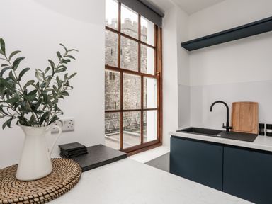 A kitchen corner with a sink cutting board plant in a white jug and a window showing a stone castle at Llety Pen Deitsh 1 in Caernarfon