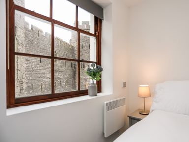 A bedroom with a bed lamp and flowers on a window sill overlooking a stone castle at Llety Pen Deitsh 1 in Caernarfon