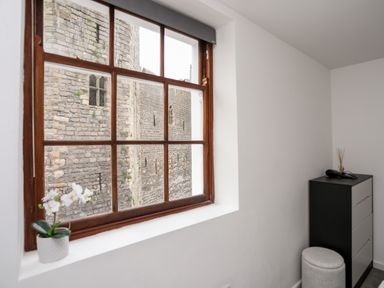 A window with brown wooden frame overlooking a stone building with a small plant on the windowsill and a black and white chest of drawers with a stool beside it at Llety Pen Deitsh 1 in Caernarfon