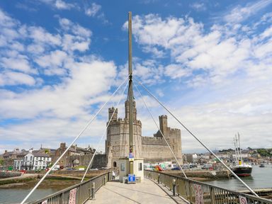 A stone castle with towers at the end of a pedestrian bridge with railing and signs over water at Llety Pen Deitsh 1 in Caernarfon