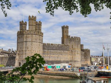 A stone castle with multiple towers near a water body with cars and people around in Caernarfon