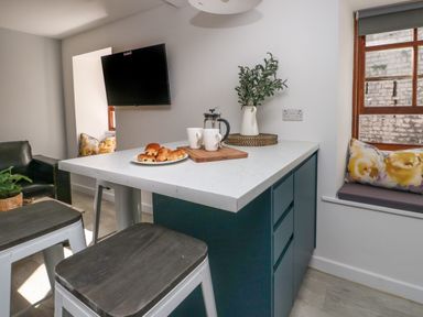 A kitchen island with stools a plate of pastries and a coffee press next to a window seat with cushions at Llety Pen Deitsh 2 in Caernarfon