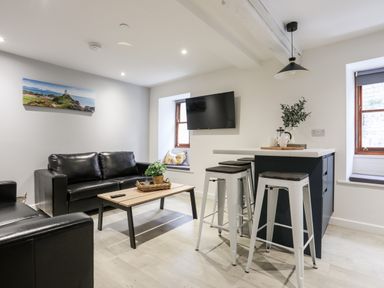 A living room with black leather sofas a wooden coffee table a wall-mounted TV a kitchen island with three bar stools and two windows at Llety Pen Deitsh 2 in Caernarfon