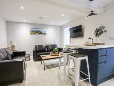 A living room with two black leather sofas a wooden coffee table a kitchen island with stools and a wall-mounted TV at Llety Pen Deitsh 2 in Caernarfon