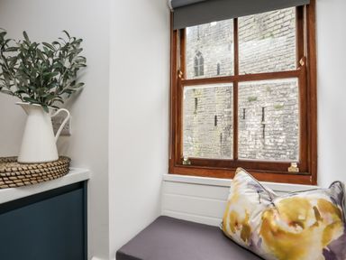 A window with a wooden frame overlooking a stone building a cushioned seat with a floral pillow and a white pitcher with green leaves on a countertop at Llety Pen Deitsh 2 in Caernarfon