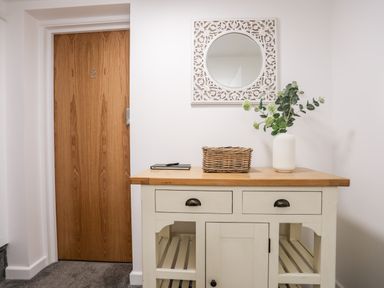 A hallway with a wooden door numbered 2 a white cabinet with a wicker basket and a vase with green plants and a decorative white framed mirror on the wall at Llety Pen Deitsh 2 Caernarfon