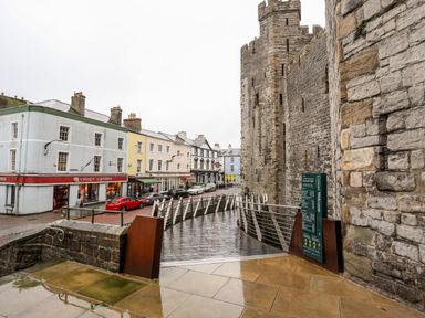A street view with a stone castle wall and a modern bridge next to pastel colored buildings and parked cars at Llety Pen Deitsh 2 in Caernarfon