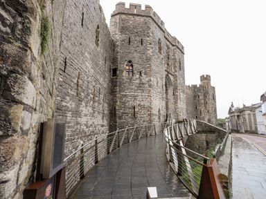 A stone castle wall with arched windows and a curved metal railing walkway beside a street with buildings at Llety Pen Deitsh 2 in Caernarfon