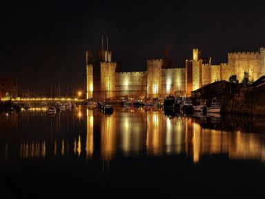 A castle illuminated at night with boats docked in front reflecting on the water at Llety Pen Deitsh 2 in Caernarfon