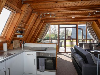A kitchen area with stove, oven, and utensils next to a living room with grey sofas and a glass door leading outside at 171 Snowdonia View in Chwilog